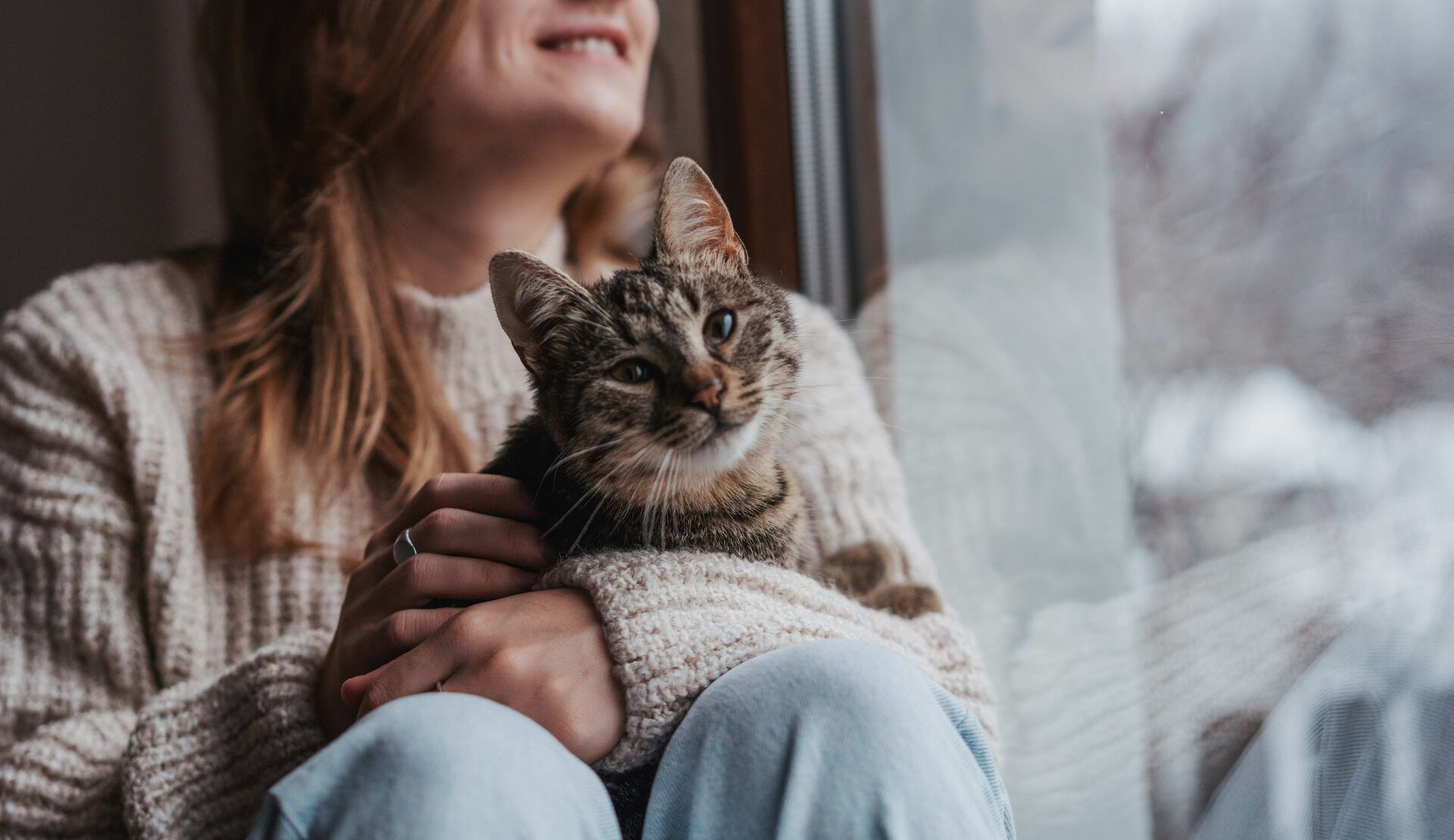 Eine Katze kuschelt mit ihrer Halterin vor einem großen Fenster.